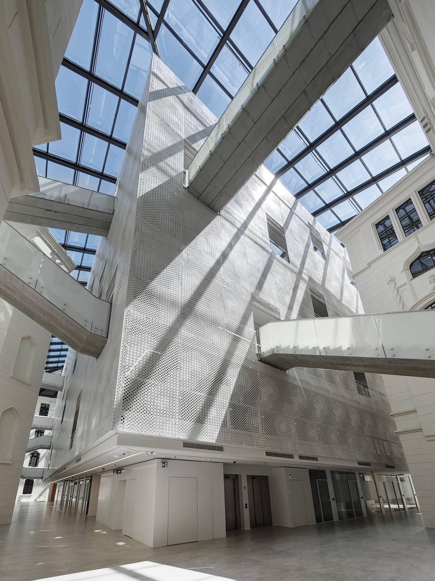 The roofed courtyard seen from below: a volume clad in perforated white metal, glazed roof structure, bridges between levels