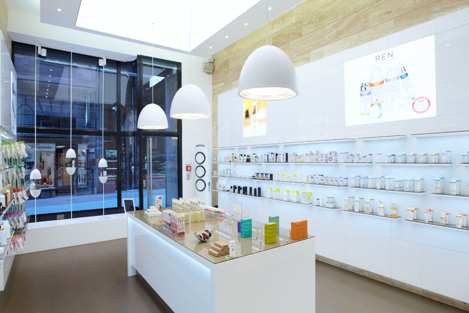 A run of backlit shelves with glass shelf plates, filled with cosmetics, next to a band of travertine